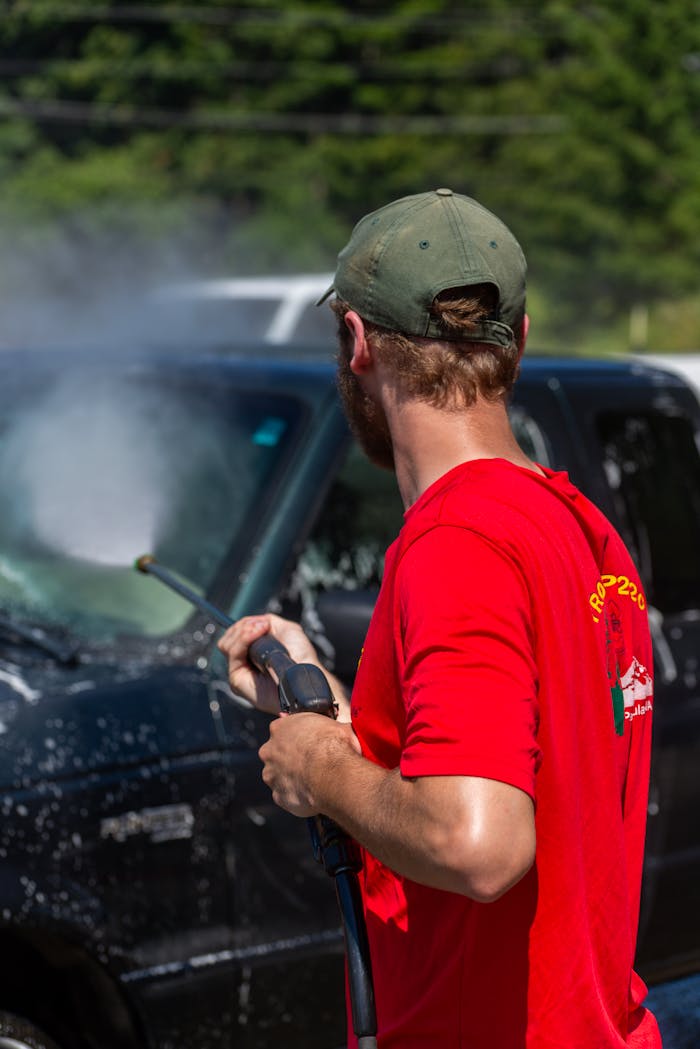 A man in a red shirt uses a power spray to clean a black car outdoors on a sunny day.